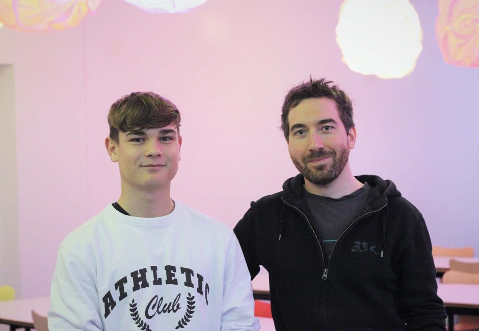 Niculin und Pascal befinden sich in der Cafeteria von Ergon. Hinter ihnen sind Tische, verschiedenfarbige Stühle und wolkenförmige Lampen zu sehen, die von der Decke hängen. Niculin und Pascal befinden sich in der Cafeteria von Ergon. Hinter ihnen sind Tische, verschiedenfarbige Stühle und wolkenförmige Lampen zu sehen, die von der Decke hängen.